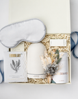 A curated gift box set on a white background, featuring a white sleep mask, a white diffuser, a white candle, a card from Formulary, and a small soap, all arranged in a cream gift box with shredded paper and blue ribbon accents.