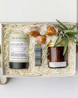 A white gift box with ivory paper shred and a green ribbon on a white background. Inside the box are a small live air plant with a care card, a mini tube of artisan jasmine green tea, a 4oz hand-poured soy candle in the scent Forest Chai, a matchbox of navy and green plaid-patterned matches, and three loose caramels — a thoughtful “Sending Thanks” gift box.