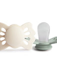 The image shows two baby pacifiers with butterfly-shaped shields, one cream-colored and the other a muted green, set against a clean white background.