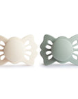 The image shows two baby pacifiers with butterfly-shaped shields, one cream-colored and the other a muted green, set against a clean white background.