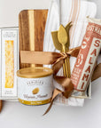 A white gift box with ivory paper shred and a brown ribbon on a white background. Inside the box are an artisan pork all-natural uncured smoked salami, everything spice flatbread crackers, a 9oz jar of sea salt and black pepper peanuts, a neutral striped tea towel, a set of three gold-plated cheese knives, a walnut charcuterie board (optionally engraved), and a small dried bunny tail decoration — an elegant gourmet charcuterie gift set.