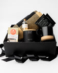 A curated gift box set on a white background, featuring a black gift box with a black ribbon, a black mug with a wooden coaster, a black candle, a bottle of whiskey, chocolate chip cookies, and a black-and-gold plaid blanket.