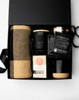 An open black gift box on a white background containing a beige-and-brown plaid blanket, a black candle, a small bottle, a black coffee bag labeled Pour Over Coffee, a Lark box, and a black mug with a beige base.
