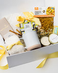 A white gift box with ivory paper shred and a yellow ribbon on a white background. Inside the box are a soft knit ivory throw blanket, chicken noodle soup mix, two lemon ginger tea sachets, two raw honey sticks, unisex plush ivory cloud socks, a 4oz hand-poured “Thinking of You” candle in the scent White Tea, honey ginger cough drops, a Moonstruck artisan milk chocolate bar, and a handcrafted dried floral mini bouquet — a deluxe “Get Well Soon” care package.