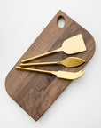 Matte gold cheese knives, set of three, gold-plated stainless steel, one pronged knife for hard cheeses, one wedge knife for semi-hard cheeses, and one tear-shaped knife for soft cheeses. Shown on top of a wooden cheese board in a white background.