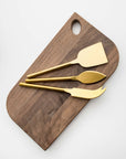 Matte gold cheese knives, set of three, gold-plated stainless steel, one pronged knife for hard cheeses, one wedge knife for semi-hard cheeses, and one tear-shaped knife for soft cheeses. Shown on top of a wooden cheese board in a white background.
