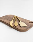 Matte gold cheese knives, set of three, gold-plated stainless steel, one pronged knife for hard cheeses, one wedge knife for semi-hard cheeses, and one tear-shaped knife for soft cheeses. Shown side way on top of a wooden cheese board in a white background.