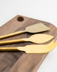 Matte gold cheese knives, set of three, gold-plated stainless steel, one pronged knife for hard cheeses, one wedge knife for semi-hard cheeses, and one tear-shaped knife for soft cheeses. Shown form a corner view on top of a wooden cheese board in a white background.