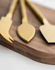 Matte gold cheese knives, set of three, gold-plated stainless steel, one pronged knife for hard cheeses, one wedge knife for semi-hard cheeses, and one tear-shaped knife for soft cheeses. Edges shown in a white background on top of a wooden cheese board.