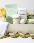 A front view of green goddess self-care gift set featuring a spa headband, a Tone face mask, a green scalp massager, a coconut milk soak, a jasmine bath bomb, a white tea trinket green jar candle, and a Nordic brush, all arranged in a white gift box with ivory shred and tied with a green ribbon on a white background.