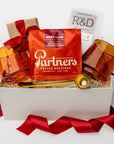A white gift box on a white background, filled with ivory paper shred. Inside the box are: a 12 oz bag of whole‑bean coffee from Partners (with tasting notes of milk chocolate, toffee, and dried fruit), a set of two modern amber glass mugs, a gold‑metal coffee scoop that also serves as a bag clip, a Markham & Fitz browned butter chocolate bar, and dark‑chocolate sea‑salt caramels — a festive “Holiday Coffee” curated gift box.