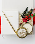 A white gift box with ivory paper shred and tied with a red ribbon on a white background. Inside the box are an ivory soft-cover pocket journal, a minimalist gold bullet pen, a 4oz hand-poured “‘Tis the Season” candle, and a handmade dried floral mini bouquet — a festive “Thankful for You” holiday gift box.