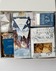 Holiday Treats Gift Box is featured with a blue ribbon crossing underneath. Contents include a German Chocolate Cake Bar, Tea Cakes (11.2 oz), S’mores Popcorn (6 oz), Snowflakes Vanilla Butter Cookies (5 oz), a 2-pack of Hot Cocoa on a Stick, Almond Toffee Treats (7.94 oz), and Sea Salt Caramel Candy (8 oz), all arranged on ivory shred. The image is displayed on a white background.