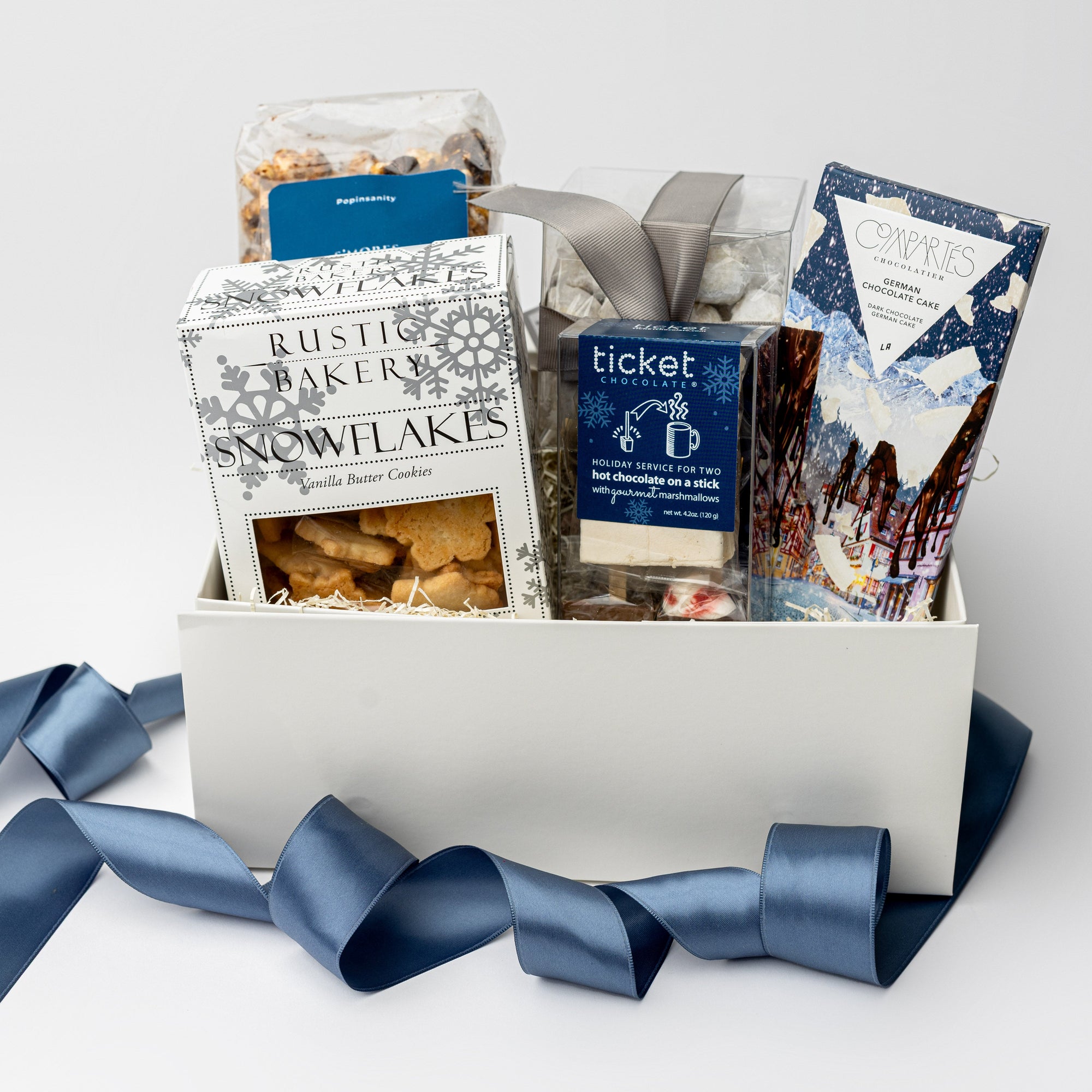 Holiday Treats Gift Box is featured with a blue ribbon crossing around it. Contents include a German Chocolate Cake Bar, Tea Cakes (11.2 oz), S’mores Popcorn (6 oz), Snowflakes Vanilla Butter Cookies (5 oz), a 2-pack of Hot Cocoa on a Stick, Almond Toffee Treats (7.94 oz), and Sea Salt Caramel Candy (8 oz), all arranged on ivory shred. The image is displayed on a white background.