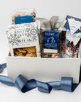 Holiday Treats Gift Box is featured with a blue ribbon crossing around it. Contents include a German Chocolate Cake Bar, Tea Cakes (11.2 oz), S’mores Popcorn (6 oz), Snowflakes Vanilla Butter Cookies (5 oz), a 2-pack of Hot Cocoa on a Stick, Almond Toffee Treats (7.94 oz), and Sea Salt Caramel Candy (8 oz), all arranged on ivory shred. The image is displayed on a white background.