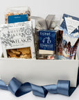 Holiday Treats Gift Box is featured with a blue ribbon crossing around it. Contents include a German Chocolate Cake Bar, Tea Cakes (11.2 oz), S’mores Popcorn (6 oz), Snowflakes Vanilla Butter Cookies (5 oz), a 2-pack of Hot Cocoa on a Stick, Almond Toffee Treats (7.94 oz), and Sea Salt Caramel Candy (8 oz), all arranged on ivory shred. The image is displayed on a white background.
