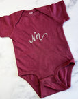 Initial embroidered onesie in rouge personalized in champagne thread with the letter M on a marble like background.