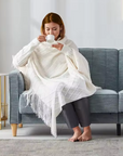 A woman wrapped in an ivory-knit throw blanket sitting on a grey couch, sipping from a white mug; the throw has a textured diamond knit pattern and fringed edges.