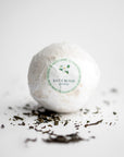 Jasmine bath bomb in its white transparent covering with white with green details label with a jasmine flower, black lettering, and with jasmine shreds around on a white background.