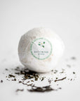 Jasmine bath bomb in its white transparent covering with white with green details label with a jasmine flower, black lettering, and with jasmine shreds around on a white background.