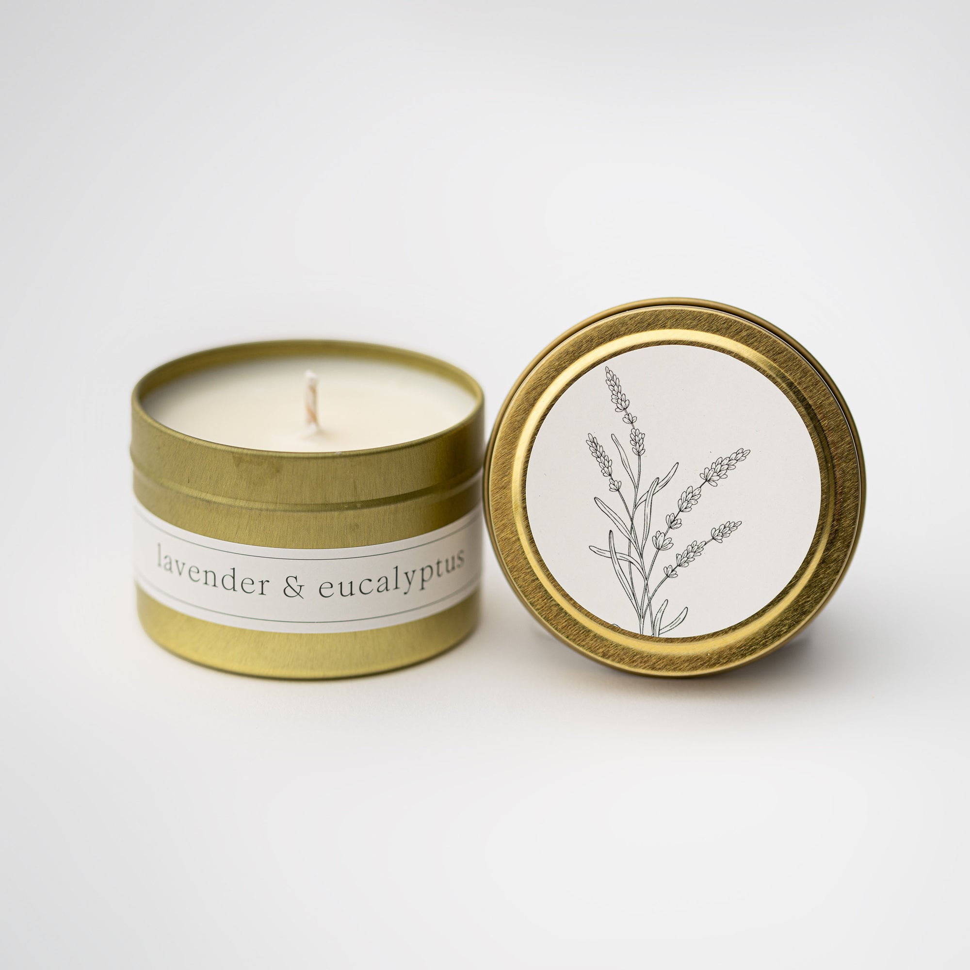 Lavender and eucalyptus travel size candle featured in a gold tin round container with white label black lettering and lid with a white round label  with a black silhouette of a lavender flower on a white background.