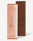 The image showcases two rectangular bars of chocolate, one wrapped and one unwrapped. The unwrapped chocolate bar displays a rich, brown hue with visible rectangular indentations, typical of chocolate bars. The wrapped bar is in a package with a soft peach-colored gradient, likely representing the flavor profile. The package features text indicating "Milk Chocolate Caramel 32%" and "Le Belge Napa Valley." The bars are clean, well-defined, and present a premium appearance.