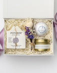 A top view of our lovely lavender gift box, a lavender-themed gift set featuring a lavender bath bomb, a 4 oz. hand-poured lavender candle, French lavender soap, and a handmade dried floral mini bouquet, all arranged in a white gift box with ivory shred and tied with a purple ribbon on a white background