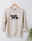 Mama bear sweatshirt featured in sand color engraved with bear silhouette with mama cut out on the middle of the image hanged in a white background with a plant in the bottom right corner.