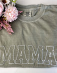 Mama embroidered comfort color tee in bay color with mama embroidered in white thread. With a pink flower on the top left part on a white background.