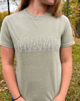A mother wearing the embroidered comfort color tee in bay color with mama embroidered in white thread. on a wood nature background.