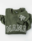 Folded army green crewneck sweatshirt with the word ‘MAMA’ printed in large white letters across the chest and a cuffed sleeve visible with the words Max, Axel and a heart emoji on a white background.