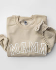 Folded nude crewneck sweatshirt with the word ‘MAMA’ printed in large white letters across the chest and a cuffed sleeve visible.
