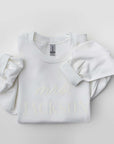 Folded white crewneck sweatshirt with the word ‘MAMA’ printed in large white letters across the chest and a cuffed sleeve visible.