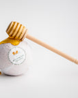Milk and Honey Bath Bomb is featured in its white, transparent wrapping with a white label that has a honeycomb image and black lettering. On top of it a honey drizzle with honey dripping on a white background.