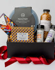 The movie night gift box is featured. A popcorn popper, a box of movie trivia, a bottle of popcorn kernels, two popcorn seasonings, and a Compartés drive-in popcorn chocolate bar are arranged inside a black gift box with brown shred, with a red ribbon draped around the base. The items are neatly placed, taller elements like the popcorn popper and popcorn bottle in the back, while the trivia box sits centrally, and the seasonings are positioned in front. On a white background.