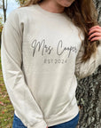 Mrs. established a custom sweatshirt in sand color in black lettering personalized with Mrs. Cooper Est. 2024, worn by a bride in wood -wood-nature-like like environment.