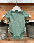 Name embroidered onesie featured in basil color, personalized in champagne thread with the name Jameson on a wooden background with two silver buckets on each side of it.