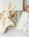 A top view of a New Baby Gift Box featuring a Baby’s First Year book, a wooden teether, a soft bunny lovey, Dandelion natural tallow balm, a handcrafted natural floral bundle, and a sage green swaddle, all arranged in a white gift box with ivory shred and tied with a blush ribbon on a white background.
