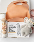 New baby girl gift box, A top view of a Baby Girl Gift Box featuring a knitted cute elephant holding roses, a floral swaddle, a wooden rattle, the book Guess How Much I Love You, and a pink silicone bib with an engraved name, all arranged in a white gift box with ivory shred and tied with a pink ribbon on a white background.