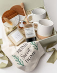 A top view of the New Home Housewarming Gift Box featuring a set of two marble and wood coasters, a “Home Sweet Home” tea towel, a 9 oz. hand-poured candle labeled “New Beginnings, New Home, New Memories” (optionally personalized with a family name), a mini tea tube of jasmine green tea, a mini jar of honey with a wooden honey drizzler, and a set of two speckled stoneware mugs, all arranged in a white gift box with ivory shred and tied with a green ribbon on a white background.