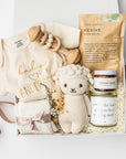 A white gift box with ivory paper shred and a brown ribbon on a white background. Inside the box are an 8-oz hand-poured lavender soy candle labeled “Amazing Mother,” a pair of cozy cloud plush socks, a Gerber onesie with personalized embroidery, Dandelion Naturals tallow balm, a Cuddle + Kind knit lamb, a wooden rattle, Slow North “Revive” natural bath salts, and a handcrafted dried floral mini bouquet — a “New Mom & Baby” gift box.