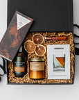 Nightcap Whiskey Cocktail Curated Gift Box, A top view of the Night Cap Gift Box presented in a black gift box filled with brown shred and tied with a black ribbon, featuring a Corkcicle whiskey glass with ice wedge, a Compartes Nightcap dark chocolate bar, scotch-infused wooden toothpicks, a charred oak & maple syrup artisan cocktail mix, a PF Candle Co. 7.2 oz Teakwood & Tobacco candle, and a crispy orange slices snack pack, all arranged on a white background.