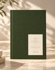 A dark green hardcover notebook with a cream paper label on the front, standing on a light marble surface with warm sunlight and leaf shadows.