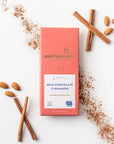 Nutty milk chocolate cinnamon bar is featured in its pink rectangular-shaped package that has gold specks, gold lettering and a label in white printed on it using brown and blue lettering. Cinnamon powder, sticks and six almonds around it on a white background.