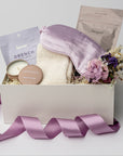 A front view of a relaxing self-care gift set presented in a white gift box filled with ivory shred and tied with a lavender ribbon, featuring a satin sleep mask, a 2 oz. Santal Rock Rose candle, a Drench sleep mask, an 8 oz. lavender oat bath soak, cloud socks, and a handmade dried floral mini bouquet, all arranged on a white background.