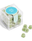 Sugarfina pear Italian ice cube is featured in its transparent square shaped container with its blue and gold details label and white lettering, revealing the green round gummies inside and in front of it 5 loose gummies all of this on a white background.