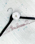 A black wooden clothes hanger is embellished with a white fabric flower and pink aluminum wire script that spells "The Dress," scattered with decorative beads.