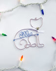 Personalized cat ornament, image shows the cat ornament done in rose gold wire that forms the outline of a cat's body and head with a white bow and a tiny silver bell. In the center of the ornament is personalized with ice blue colored wire with the name Ellie.