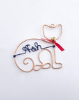 Personalized cat ornament, featuring an image of a cat crafted from gold aluminum wire that forms the outline of a cat's body and head, complete with a red bow and a sgold bell. In the center is personalized with the name Ash in black aluminum wire.