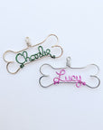 Personalized dog ornament,dog-bone-shaped outline, one in gold wire aluminum with the name charlie in the center in green and the other one in silever color aluminum wore in the center the name lucy in pink .